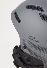 Gray helmet with ventilation slots, textured accents, and adjustable straps. Features branding "Sweet Protection" on the side.