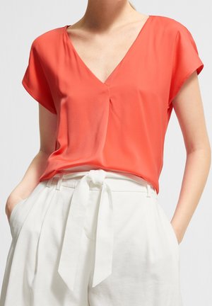 Kobieta w koralowej bluzce z krótkim rękawem i dekoltem w serek, włożonej w białe, wysokowodne spodnie z materiałowym paskiem związanym w kokardę i rękami w kieszeniach.