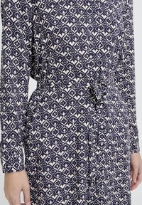 Jūrininkų ir baltos spalvos geometrinio rašto ilgos rankovės suknelė, su mygtukais priekyje ir surišta juosta, demonstruojama ant asmens liemens ir rankų.