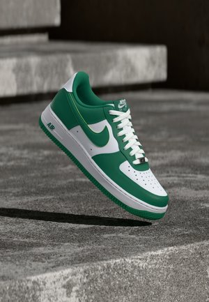 Grüner und weißer Nike Air Force 1 Sneaker schwebt über einer Betonsurface mit Stufen im Hintergrund.