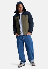 Polstret jakke i olivengrøn og marineblå, med hætte, lynlåslukning og elastiske manchetter. Kombineret med løstsiddende blå jeans og grå sneakers.
