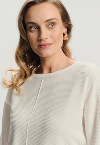Kobieta z falującymi blond włosami, nosząca duże złote geometryczne kolczyki oraz strukturalną, kremową bluzkę, patrząca nieco w prawo z neutralnym wyrazem twarzy.