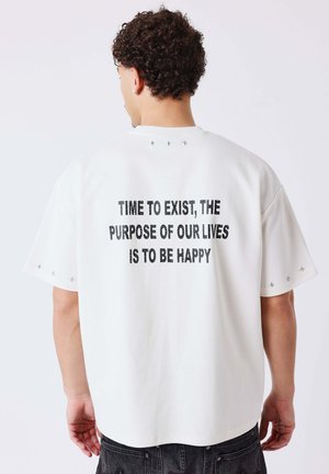 Man met krullend haar die achterwaarts kijkt, draagt een wit t-shirt met de tekst "Tijd om te bestaan, het doel van ons leven is gelukkig zijn".