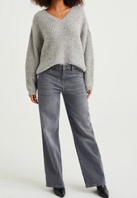 Grauer Strickpullover mit V-Ausschnitt und übergroßen Ärmeln, kombiniert mit weit geschnittenen grauen Jeans. Schwarze Absatzschuhe vervollständigen das Outfit.