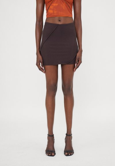 Jupe noire mini avec un design ajusté, présentant un drapé asymétrique d'un côté et une texture lisse. Assortie à des talons noirs à lanières.