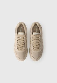 Sneakers bege com materiais em malha e camurça, apresentando atacadores planos, um colar acolchoado e detalhes em dourado nas laterais. Sola de borracha texturizada.