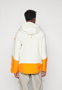 White and orange waterproof jacket with a hood, featuring a logo on the back, orange accents at the hem and sleeves, and a relaxed fit.