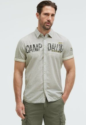 Mann mit kurzen braunen Haaren, der ein hellgraues kurzärmeliges Hemd mit Knöpfen und dem Text "CAMP DAVID" sowie olivgrüne Cargohosen trägt.
