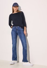 Marineblauwe langesleeve top, lichtblauwe uitlopende jeans en witte sneakers. Pet in bijpassende marineblauwe tint. Gladde textuur, aansluitend ontwerp en denim detail.
