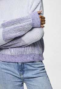 Lila pletena pulover s dugim rukavima, ukrašen vertikalnim prugama i mekom teksturom, uparen s plavim trapericama.