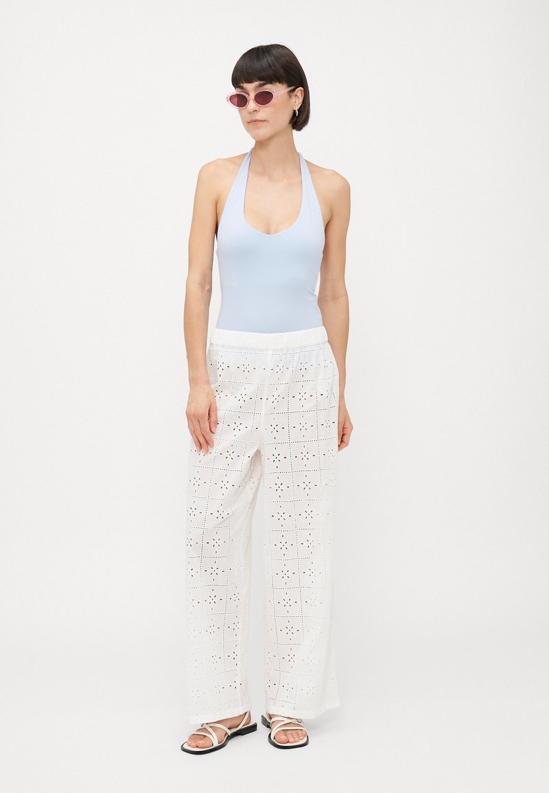 Vrouw in een lichtblauwe haltertop, witte wijde oogjesbroek, roze cat-eye zonnebril en witte sandalen die tegen een effen achtergrond staat.