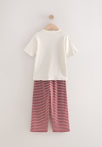 Hvid T-shirt med korte ærmer og rund hals udstyret med røde og hvide stribede bukser hængende på en træbøjle mod en neutral baggrund.