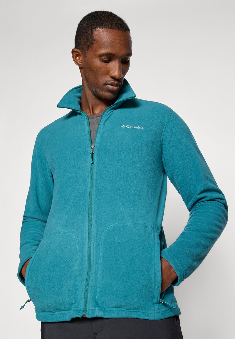 Zielona polarowa kurtka z zamkiem błyskawicznym i wysokim kołnierzem, z dwoma przednimi kieszeniami oraz regulowanym sznurkiem na dole, z subtelnym logo po lewej stronie klatki piersiowej.