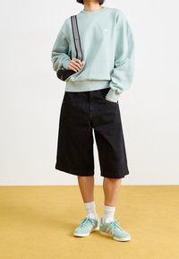 Persona vestida con un sudadera verde menta, pantalones cortos negros de pierna ancha hasta la rodilla, calcetines blancos y zapatillas azul menta de pie sobre un suelo amarillo.