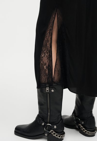 Μαύρα μποτάκια αστραγάλου με μυτερή μύτη, μεταλλικό φερμουάρ και αλυσίδα ως διακοσμητικό. Μαύρο φόρεμα με πλάγια σκίσιμο που αποκαλύπτει δαντέλα από κάτω.