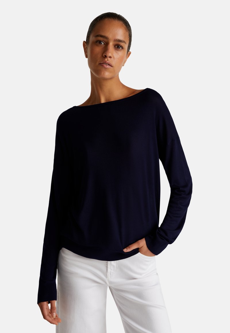 Marineblauwe gebreide top met lange mouwen, een wijde halslijn en een ontspannen fit, gecombineerd met witte jeans. Soepele textuur, casual ontwerp.