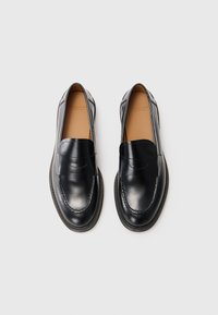Mocassins en cuir noir avec une texture lisse, un bout rond et des détails de couture. Doublure en tan doux et design minimaliste pour un enfilage facile.