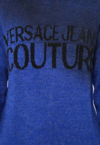 Kék kötött pulóver, amelyen a "VERSACE JEANS COUTURE" felirat nagy fekete betűkkel található a mellkason. Puha tapintású és közepes súlyú anyag.