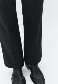 Black tailored trousers with a straight cut and visible seams, paired with black textured loafers featuring a sleek square toe.
