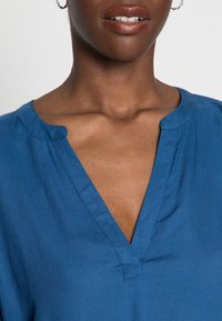 Blå bomullsskjorta med v-ringad design, med en enkel krage och slät textur. Inga påtagliga mönster eller detaljer.