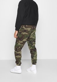 Kamouflage-mönstrade sweatpants i grön och brun, med elastiska avslut och midja, matchade med vita högsneakers. Svart sweatshirt synlig.