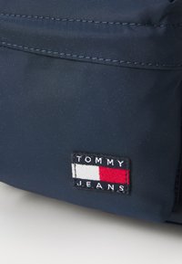 Borsa di tessuto blu navy con un evidente logo ricamato "TOMMY JEANS" con accenti rossi, bianchi e blu nella parte inferiore. Texture liscia.