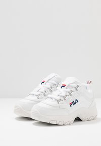 Baskets de sport blanches avec une tige texturée, dotées d'un système de laçage, d'un logo en bleu et rouge, et d'une semelle épaisse et à motifs.