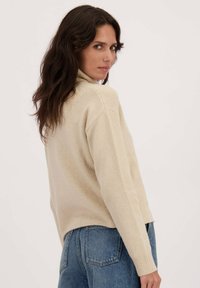 Beige oversized coltrui met een gestructureerde afwerking, met verlaagde schouders en geribbelde manchetten, gecombineerd met blauwe high-waisted jeans.