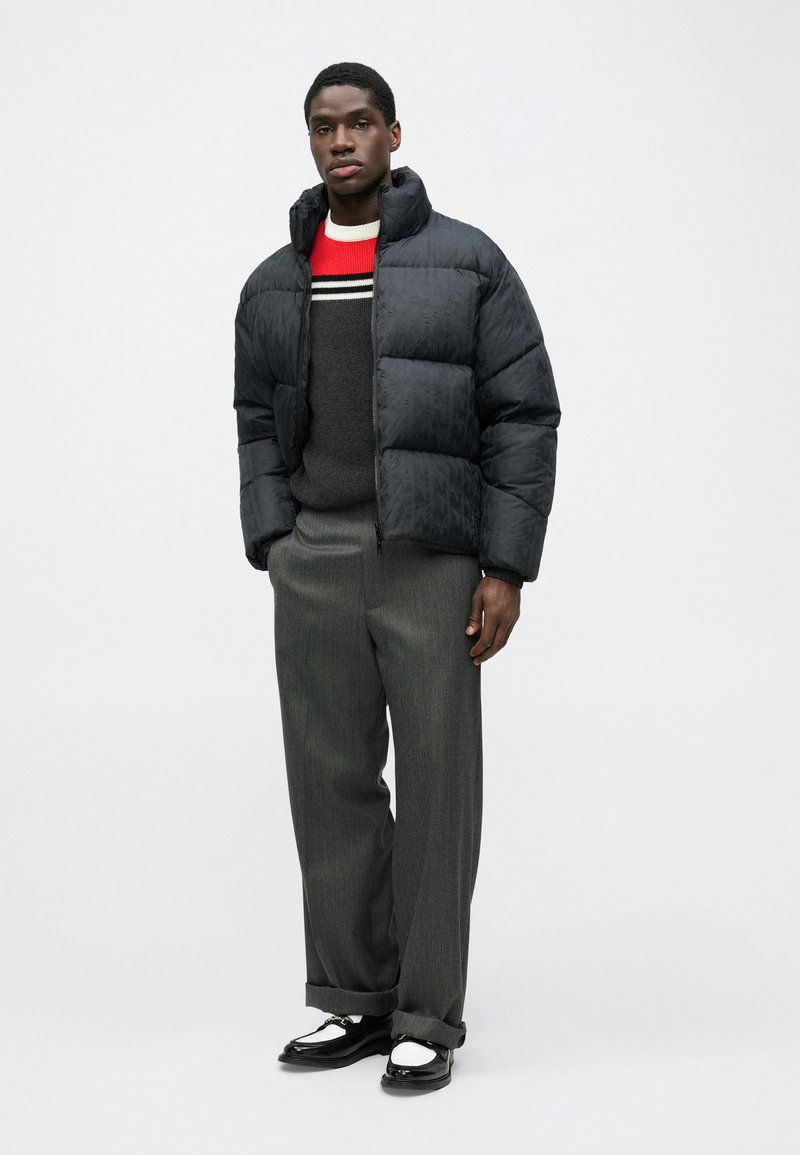 Puffer jacket in dark blue with a glossy finish, black ribbed cuffs, styled over a gray sweater with red and white stripes, gray trousers, and black shoes.