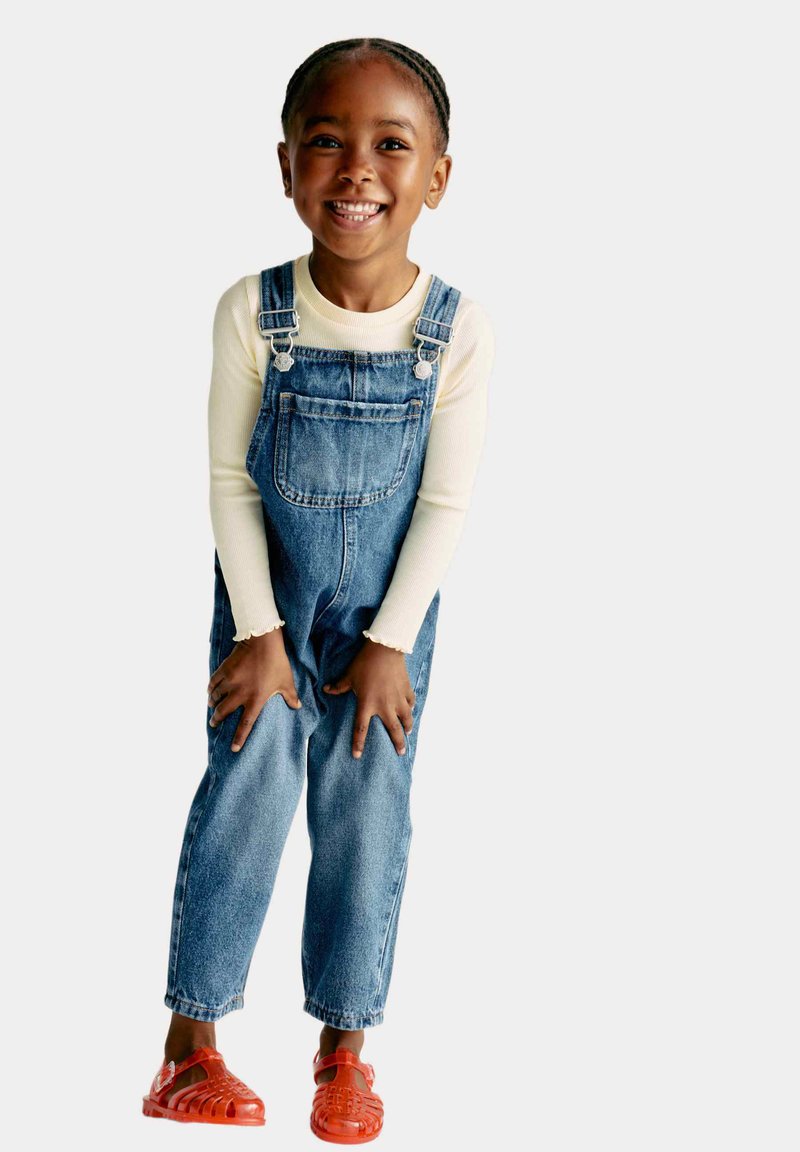 Leren denim overall in lichtblauw met verstelbare banden, een voorzak en omgeslagen broekspijpen, gecombineerd met een witte lange mouwen shirt en rode sandalen.