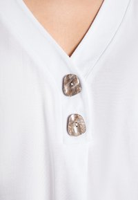 Witte blouse met V-halsontwerp en twee decoratieve schelpachtige knopen met een gladde textuur.
