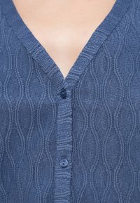 Μπλε ναυτικού χρώματος υφασμάτινο πουκάμισο με κουμπιά, με V-λαιμόκοψη, διακοσμημένο με κυματιστό μοτίβο και ένα μπλε κουμπί στο μπροστινό μέρος.