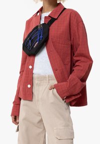 Persona que lleva una chaqueta de cuadros rojos, una camisa blanca, pantalones cargo beige y un bolso cruzado negro con detalles en cordón azul en el pecho.