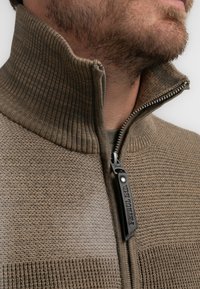 Chaqueta marrón de textura con cremallera y cuello acanalado. Presenta una cremallera lisa de contraste y una etiqueta que dice "Petrol Ind." en la cremallera.