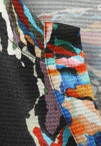 Mehrfarbiger Stoff mit komplexen Mustern in Schwarz, Blau, Orange und Grün, mit sichtbaren Nähten und strukturierten Oberflächendetails.