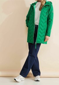 Groene gewatteerde jas met een chevronpatroon, verstelbare capuchon en ritssluiting aan de voorkant. Gecombineerd met donkere denim jeans en witte sneakers.