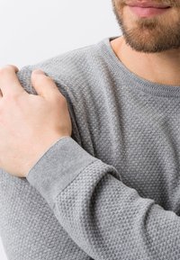 Grauer strukturierter Pullover mit einem gestrickten Muster, rundem Ausschnitt und gerippten Bündchen, dargestellt mit einer Hand, die auf dem Schulterbereich ruht.