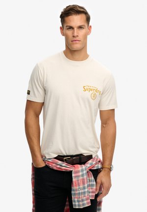 Hombre con camiseta beige de Superdry, pantalones negros, camisa de cuadros atada a la cintura y reloj de pulsera, de pie con las manos en los bolsillos.