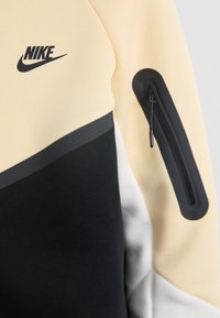 Nike sudrabs ar krēma un melnā krāsā bloku dizainu, ar faktūrētu rāvējslēdzēja kabatu uz piedurknes un gludu auduma apdari.