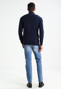 Marinblå långärmad tröja, med ribbade muddar och nederkant, i kombination med ljusblå jeans och svarta skor, visad bakifrån mot en ljus bakgrund.