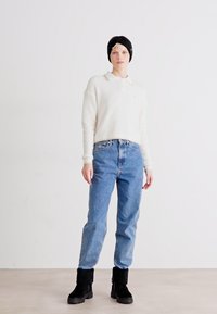 Maglione polo a maniche lunghe color crema, jeans high-waisted blu chiaro e stivaletti neri alla caviglia. Il modello indossa una fascia nera. Sfondo neutro.