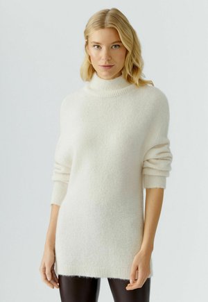 Übergroßer cremefarbener Rollkragenpullover aus weichem, flauschigem Material mit gerippten Bündchen und Saum, kombiniert mit schwarzen Leggings.