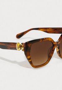 Ochelari de soare cu tortoiseshell, cu o formă îndrăzneață de cat-eye, lentile în degrade de culoare maro închis și un accent auriu pe bradă cu un detaliu hexagonal.