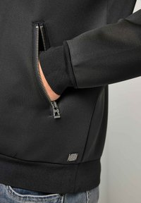 Giacca nera con cerniera e tasca laterale. Ha una texture liscia e polsini a coste, con una piccola toppa con logo vicino all'orlo.