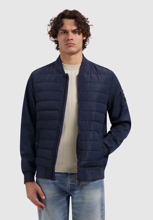 Man met krullend haar, gekleed in een marineblauwe doorgestikte bomberjack over een beige geribbelde trui en lichtblauwe spijkerbroek, staand tegen een effen achtergrond.