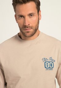 T-shirt beige a maniche lunghe in cotone con colletto a girocollo, caratterizzato da testo e numeri ricamati in blu sul petto.