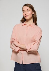 Camicia button-down rosa chiaro in tessuto morbido, dotata di colletto, maniche lunghe arrotolate fino ai gomiti e vestibilità rilassata.