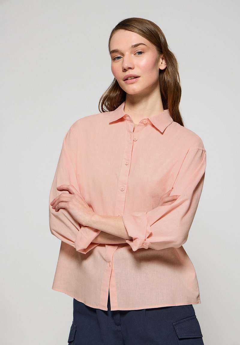 Camicia button-down rosa chiaro in tessuto morbido, dotata di colletto, maniche lunghe arrotolate fino ai gomiti e vestibilità rilassata.