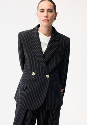 Vrouw gekleed in een zwart krijtstreep dubbelbreasted blazer met gouden knopen en bijpassende broek, met een wit overhemd en gouden oorringen.