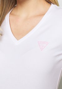 White v-neck t-shirt made of soft cotton, featuring a pink triangular logo on the left chest area. Smooth texture with fitted design.